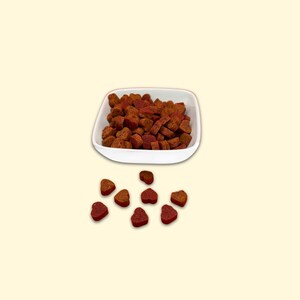 May include: A white bowl filled with heart-shaped brown and red dog treats. Some treats are scattered on a light beige surface.