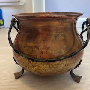 Puede incluir: Un caldero redondo de cobre con un borde ancho, que cuenta con un asa giratoria de metal oscuro unida a los lados y tres pequeños pies de metal curvados en la base. La superficie de cobre muestra una pátina desgastada con tonos naranjas y marrones.