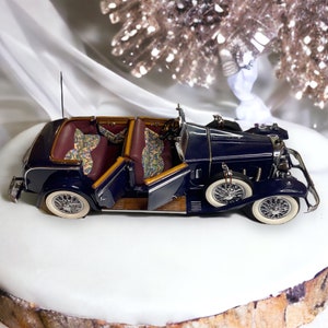 May include: A dark blue vintage car model with gold accents and a patterned interior. The car is displayed on a white cake stand with a wood base.