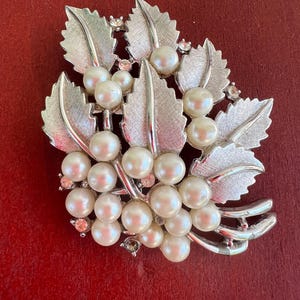 Può includere: Una spilla color argento con un grappolo di perle finte bianche, circondato da foglie d'argento testurizzate e piccoli strass trasparenti. Il metallo ha una finitura spazzolata e il design è modellato in un motivo botanico.