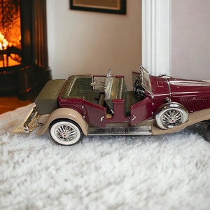 May include: A burgundy and beige vintage car model with open doors, sitting on a white fluffy surface. The car has chrome accents and a detailed interior.