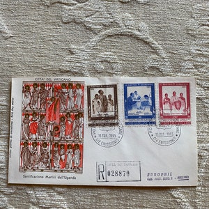 May include: A white envelope with a red and blue stamp featuring a painting of a group of people. The envelope has a red postmark that reads "16 MAR. 1965" and "DIE EMISSIONIS". The envelope also has a blue stamp with the text "CITTA' DEL VATICANO" and "R 028870". The envelope has the text "Santificazione Martiri dell'Uganda" and "EUROPHIL CAS. POST. SUCC. 1 BOLZANO".