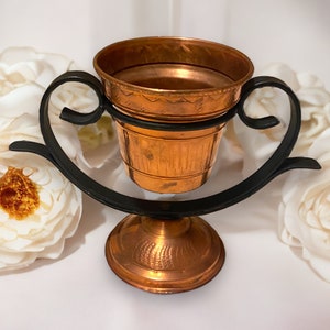 Könnte beinhalten: Ein Kupfertopf mit einem schwarzen Metallständer. Der Topf hat ein dekoratives Design und ist perfekt für die Präsentation kleiner Pflanzen oder Blumen.