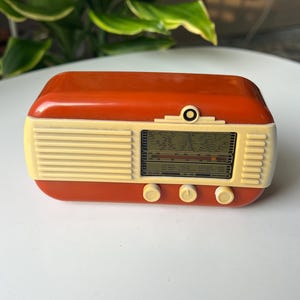 Puede incluir: Una radio de sobremesa rectangular con carcasa de plástico naranja brillante y panel frontal de color crema. La parte delantera presenta una rejilla de altavoz horizontal, una pantalla central de sintonización AM y FM y tres mandos circulares.