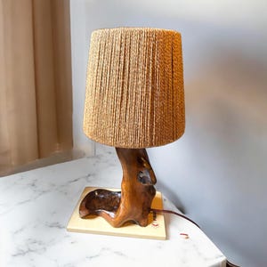 Op de afbeelding: Een tafellamp met een basis van natuurlijk hout en een geweven, beige lampenkap. De basis is gevormd als een tak en de lampenkap is cilindrisch.