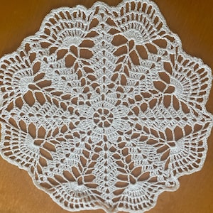 Könnte beinhalten: Ein weißes, kreisförmiges Deckchen mit einem komplizierten Häkelmuster. Das Design zeigt ein zentrales Blumenmotiv, das von strahlenförmigen, fächerartigen Elementen und einem gewellten Rand umgeben ist. Das handgefertigte Textil wird auf einer Holzoberfläche präsentiert.