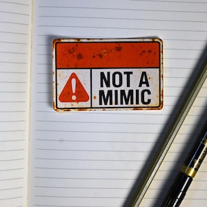 May include: A rectangular sign with a rusty orange top and white bottom, featuring a red triangle with an exclamation point and the words "NOT A MIMIC" in black. The sign is placed on a lined notebook page.