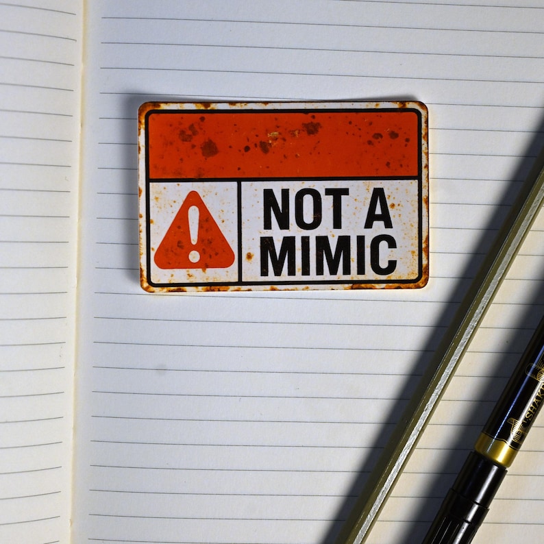 May include: A rectangular sign with a rusty orange top and white bottom, featuring a red triangle with an exclamation point and the words "NOT A MIMIC" in black. The sign is placed on a lined notebook page.