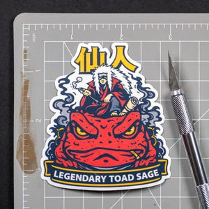Puede incluir: Una pegatina que representa a un personaje con una t&uacute;nica roja y pelo blanco, sentado sobre un sapo rojo. La pegatina tiene el texto "Legendary Toad Sage" y caracteres japoneses. Un c&uacute;ter plateado est&aacute; al lado de la pegatina sobre una esterilla de corte gris.