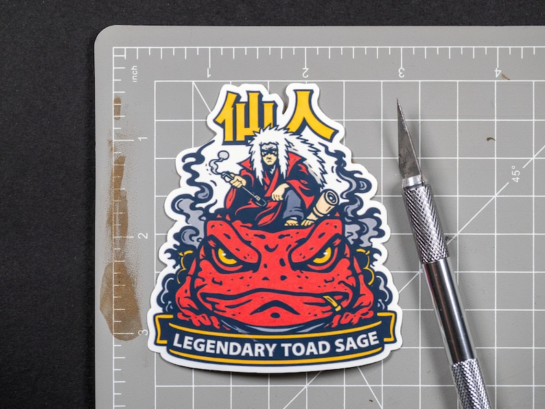 Puede incluir: Una pegatina que representa a un personaje con una t&uacute;nica roja y pelo blanco, sentado sobre un sapo rojo. La pegatina tiene el texto "Legendary Toad Sage" y caracteres japoneses. Un c&uacute;ter plateado est&aacute; al lado de la pegatina sobre una esterilla de corte gris.
