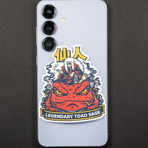 Puede incluir: Una pegatina con una ilustraci&oacute;n de dibujos animados de un personaje con cabello blanco y una t&uacute;nica roja, fumando un cigarrillo, sentado sobre un sapo rojo. El texto "Legendary Toad Sage" est&aacute; en una pancarta debajo.