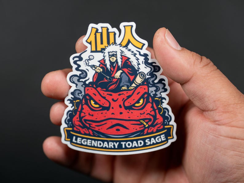 Puede incluir: Una pegatina con una ilustraci&oacute;n de dibujos animados de un personaje con cabello blanco y una t&uacute;nica roja, sentado sobre un sapo rojo. La pegatina tiene el texto "Legendary Toad Sage" y caracteres japoneses.