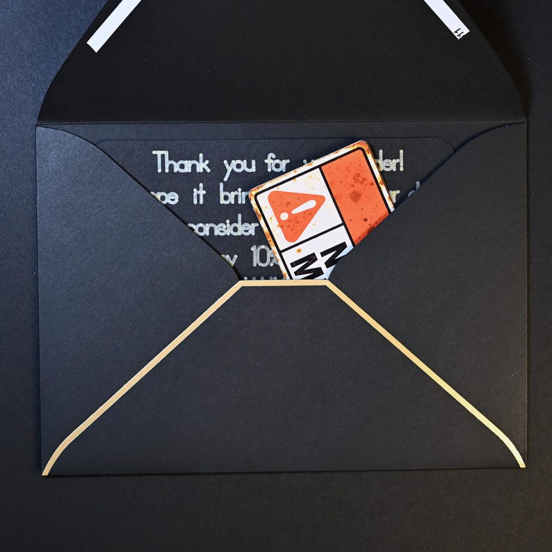 May include: A black envelope with a gold trim and a white stripe on the flap. Inside, a card with the text "Thank you for..." and a small, rectangular sticker with an orange and white design is visible.