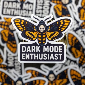May include: A sticker featuring a moth design with a skull on its head, in shades of black, yellow, and orange. The sticker has the text "DARK MODE ENTHUSIAST" in white letters on a black background. Other similar stickers are in the background.