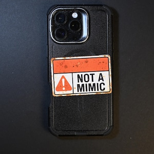 May include: Black phone case with a rectangular sticker. The sticker has a white background with an orange border and the words "NOT A MIMIC". An exclamation point is inside a triangle on the left side of the sticker.