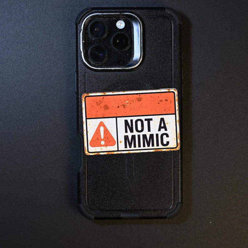 May include: Black phone case with a rectangular sticker. The sticker has a white background with an orange border and the words "NOT A MIMIC". An exclamation point is inside a triangle on the left side of the sticker.