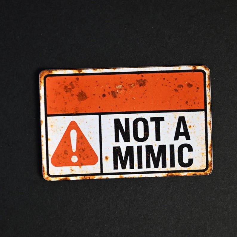 May include: Rectangular metal sign with an orange top section and white bottom section. The sign features the text "NOT A MIMIC" in black, with an orange warning symbol. The sign has a distressed, rusty appearance.
