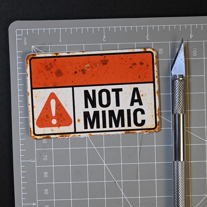 May include: A rectangular sign with a rusty orange top section and the text "NOT A MIMIC" in bold black letters. The sign has a white bottom section with an orange exclamation point symbol. A silver utility knife is next to the sign.