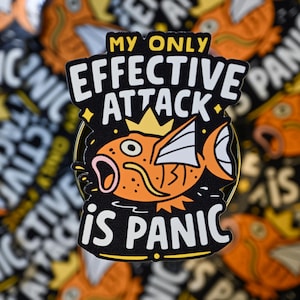May include: A black and orange enamel pin featuring a cartoon fish with a crown. The pin reads "MY ONLY EFFECTIVE ATTACK IS PANIC" in white text. The fish is orange with white fins and a pink mouth.