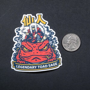 Puede incluir: Una pegatina con una ilustraci&oacute;n de dibujos animados de una figura con una t&uacute;nica roja sentada sobre un sapo rojo. La pegatina tiene el texto "Legendary Toad Sage" y caracteres japoneses. Hay una moneda al lado.