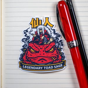 Puede incluir: Una pegatina con un sapo rojo con ojos amarillos y un personaje fumando en pipa. El personaje lleva una t&uacute;nica roja y tiene el pelo blanco. La pegatina tiene el texto "Legendary Toad Sage" y caracteres japoneses.