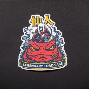 Puede incluir: Una pegatina con una ilustraci&oacute;n de dibujos animados de una figura con pelo blanco y una t&uacute;nica roja, fumando en pipa, sentado sobre un gran sapo rojo. El texto "Legendary Toad Sage" est&aacute; en una pancarta, con caracteres japoneses encima.