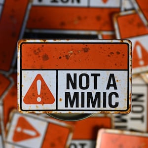 May include: A rectangular metal sign with an orange top and a white bottom section. The sign features a red triangle with an exclamation point and the words "NOT A MIMIC" in black. The sign has a rusty, distressed appearance.