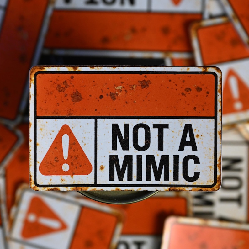 May include: A rectangular metal sign with an orange top and a white bottom section. The sign features a red triangle with an exclamation point and the words "NOT A MIMIC" in black. The sign has a rusty, distressed appearance.