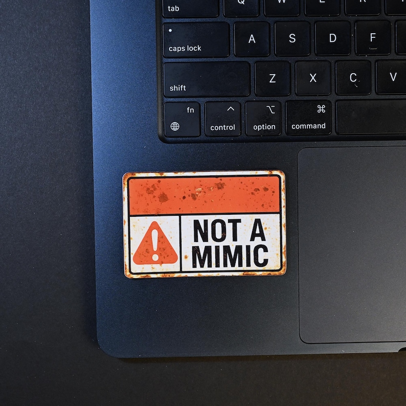 May include: A rectangular sticker with a distressed orange and white design. The sticker features the text "NOT A MIMIC" in bold white letters, with an exclamation point symbol. The sticker is placed on a laptop.