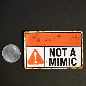May include: A rectangular sign with an orange and white design, featuring the text "NOT A MIMIC" in bold black letters. The sign has a rusty, distressed appearance. A US quarter is next to the sign for size comparison.