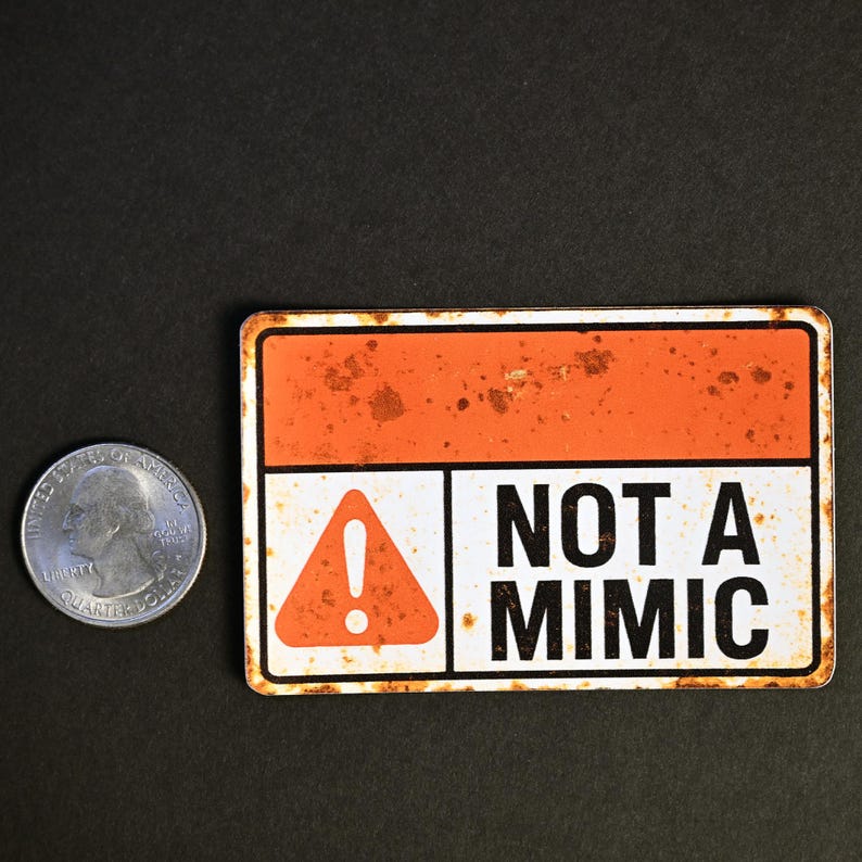 May include: A rectangular sign with an orange and white design, featuring the text "NOT A MIMIC" in bold black letters. The sign has a rusty, distressed appearance. A US quarter is next to the sign for size comparison.