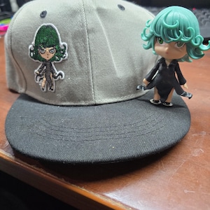 Peut inclure: Une casquette de baseball grise et noire avec un personnage d'anime brodé en vert et noir sur le devant. Une figurine de personnage d'anime verte et noire est assise sur le bord de la casquette.