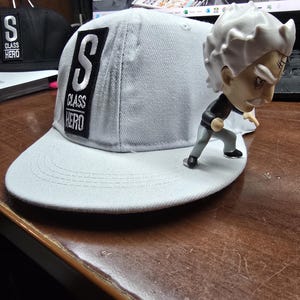 Peut inclure: Une casquette de baseball grise avec un patch noir qui dit "S CLASS HERO" en lettres blanches. La casquette est posée sur une surface en bois avec une petite figurine blanche d'une personne aux cheveux hérissés.