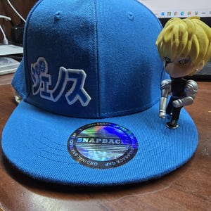 Peut inclure: Une casquette de baseball bleue avec un logo blanc qui dit "SNAPBACK" et un design de caractères japonais blancs. La casquette est posée sur une surface en bois avec une petite figurine d'un personnage aux cheveux jaunes.