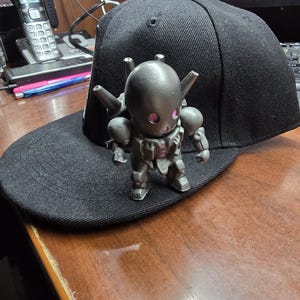 Peut inclure: Une casquette de baseball noire avec une figurine de robot argentée sur le dessus. Le robot a une tête ronde avec des yeux roses et porte une combinaison argentée avec des accents noirs.