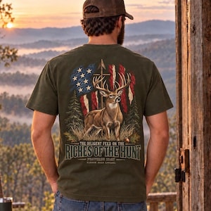May include: An olive green t-shirt featuring a graphic on the back of a deer standing in front of a distressed American flag and pine trees. The text reads: THE DILIGENT FEED ON THE RICHES OF THE HUNT PROVERBS 12:27 NARROW ROAD APPAREL.