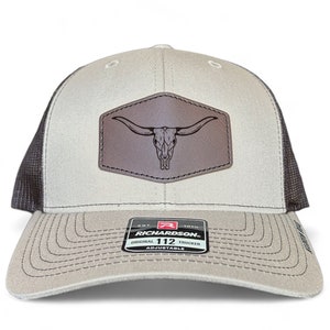 Peut inclure: Un chapeau de camionneur beige et noir avec un patch en cuir marron représentant un crâne de taureau. Le chapeau porte le texte "Richardson 112 Original Trucker Adjustable" sur l'étiquette.