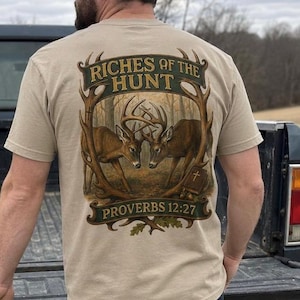 May include: A beige short-sleeve t-shirt featuring a graphic on the back with two deer facing each other, framed by antlers. The text reads "RICHES OF THE HUNT" and "PROVERBS 12:27" with an illustration of a book featuring a cross symbol.