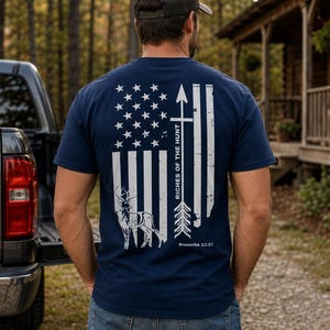 May include: A navy blue short-sleeve t-shirt featuring a white graphic on the back. The design includes a distressed American flag, a vertical arrow, a deer silhouette, and the text Riches of the Hunt and Proverbs 12:27 printed in white.
