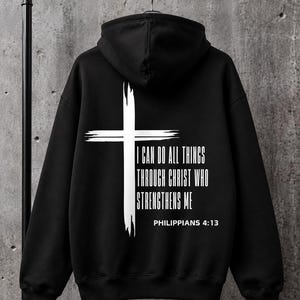 May include: A black hooded sweatshirt featuring a white graphic on the back. The design includes a distressed-style cross and the text "I CAN DO ALL THINGS THROUGH CHRIST WHO STRENGTHENS ME" followed by the citation "PHILIPPIANS 4:13" in white lettering.