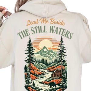 May include: A beige hoodie with a graphic print of a mountain scene with a cabin, a river, and a bear. The text "Lead Me Beside The Still Waters" is printed on the hoodie.
