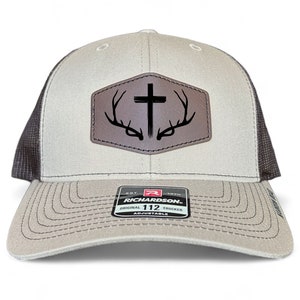 Puede incluir: Una gorra de camionero beige y negra con un parche de cuero marrón que presenta una cruz y cuernos de ciervo. El parche tiene el texto "EST. 1870 RICHARDSON 112 ORIGINAL TRUCKER ADJUSTABLE".