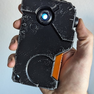 May include: A black and orange handheld device with a textured surface and a small antenna. The device has a glowing blue light and is held in a hand. The design has a futuristic, industrial aesthetic.
