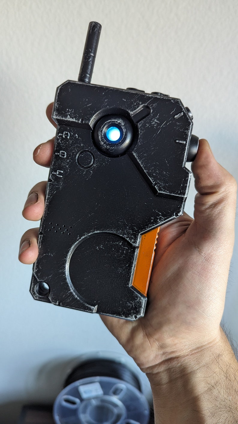 May include: A black and orange handheld device with a textured surface and a small antenna. The device has a glowing blue light and is held in a hand. The design has a futuristic, industrial aesthetic.