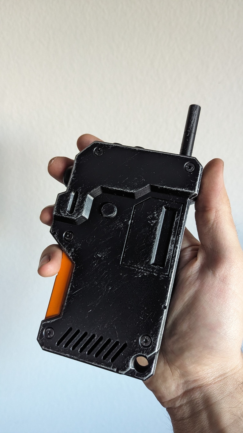 May include: A black and orange handheld device with a textured, worn appearance. The device has a rectangular shape with a small antenna and a series of screws. The orange section is on the side. The device is held in a hand.