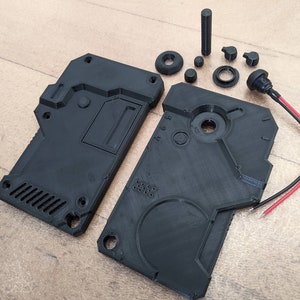 May include: Black 3D-printed electronic device components, including two rectangular housings, various circular and cylindrical parts, and a connector with red and black wires. The parts are arranged on a light brown surface.