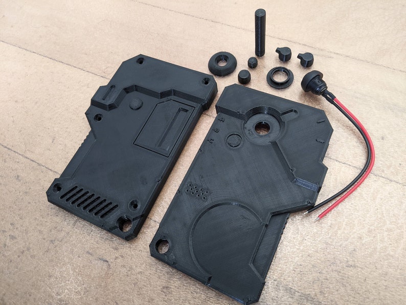 May include: Black 3D-printed electronic device components, including two rectangular housings, various circular and cylindrical parts, and a connector with red and black wires. The parts are arranged on a light brown surface.
