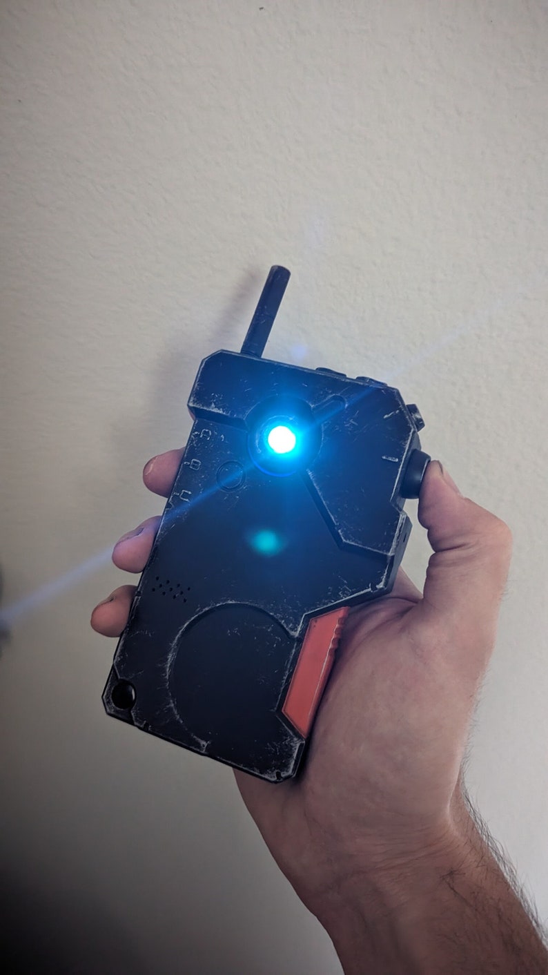 May include: A black and red device with a blue light emanating from the center. The device has a textured surface and a small antenna. The device is held in a hand.