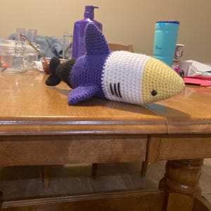 May include: A handmade crochet shark toy in shades of purple, white, yellow, and black. The shark has a pointed dorsal fin, side fins, and a tail. The shark's body is white with black gills and a yellow snout. The toy is lying on a wooden table.