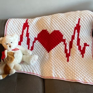 Puede incluir: Una manta blanca tejida a ganchillo con un corazón rojo y un diseño de latido cardíaco. Un oso de peluche blanco con una cinta roja descansa sobre la manta. La manta tiene un borde rojo y está sobre una superficie marrón.